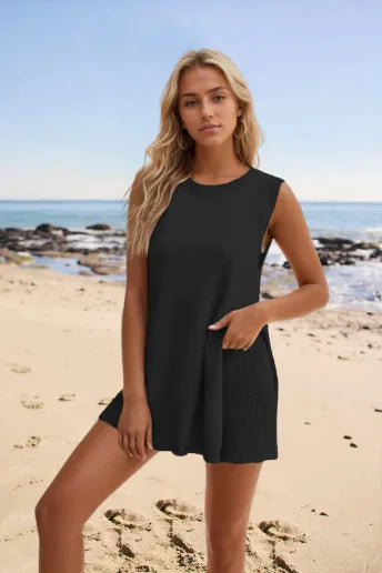 Woman standing on a sandy beach wearing a sleeveless, rust-orange mini dress with a relaxed fit and subtle ribbed texture on the sides, with the ocean and rocks in the background under a clear blue sky