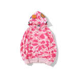 A pink and white camouflage zip-up hoodie with a full hood and ribbed cuffs, shown against a plain light gray background. The hood features bold graphic details including orange lettering and eye motifs near the top.