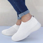 White slip-on knit sneakers with textured panels and thick white soles, shown worn with cuffed jeans.