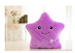 A purple star-shaped plush toy with a smiling face sits on a table in a softly lit room, with a small hanging loop at the top.