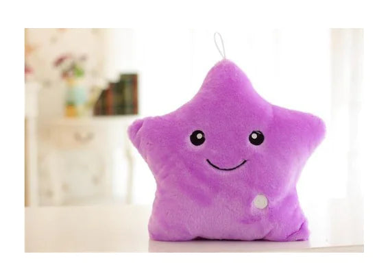 A purple star-shaped plush toy with a smiling face sits on a table in a softly lit room, with a small hanging loop at the top.