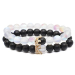 Two stretchy beaded bracelets—one with matte black beads and one with translucent white iridescent beads—linked by a small gold crown charm with rhinestones.