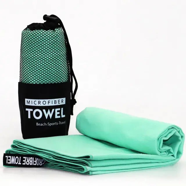 A microfiber towel in a black mesh carrying bag with the label "MICROFIBER TOWEL" on it. The towel is light green, unfolded and lying flat, showing a soft, smooth texture.