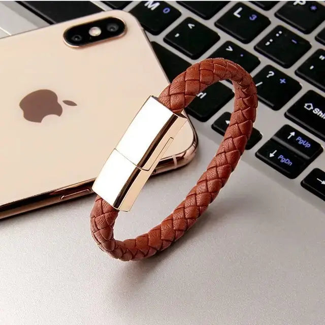 A brown braided leather bracelet with a polished metallic clasp, displayed on a silver laptop beside a gold iPhone. The bracelet has a sleek, modern design with a woven texture and reflective rectangular closure.