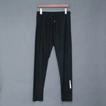 A pair of black athletic joggers hangs on a wooden hanger against a plain gray background. The joggers feature an elastic waistband with a drawstring for adjustable fit. A small white reflective design is visible near the lower leg, adding a subtle sporty touch. The fabric appears lightweight and stretchy, making the joggers suitable for casual wear, workouts, or lounging.