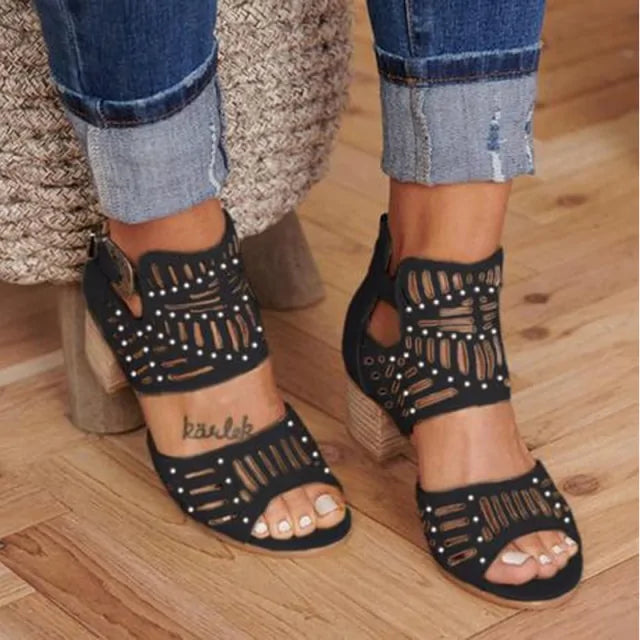 A close-up view of a person wearing stylish beige cut-out heeled sandals with ankle straps and decorative perforations, paired with cuffed blue denim jeans on a wooden floor.