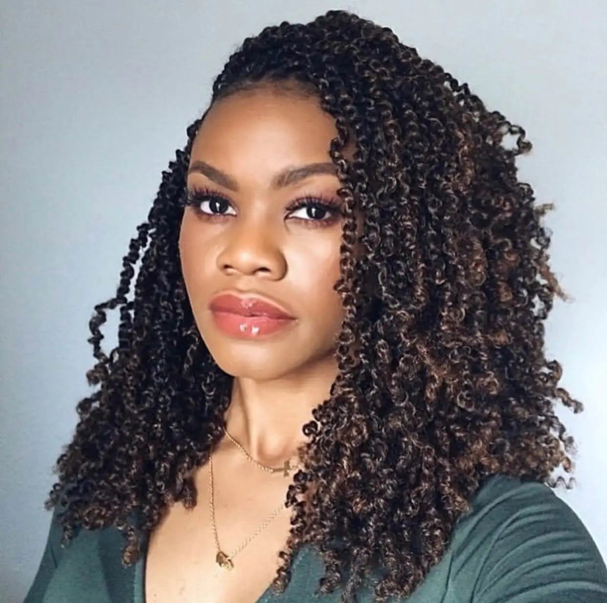 Close-up portrait of a woman with long, tight curly twists styled to one side, wearing makeup and a dark green top against a light background.