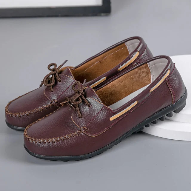 A pair of burgundy slip-on loafers with a pebbled leather-look finish, hand-stitched moc-toe detailing, and decorative lace ties. They feature side cutout accents, a soft interior lining, and a low-profile black rubber sole for a casual driving-shoe style.