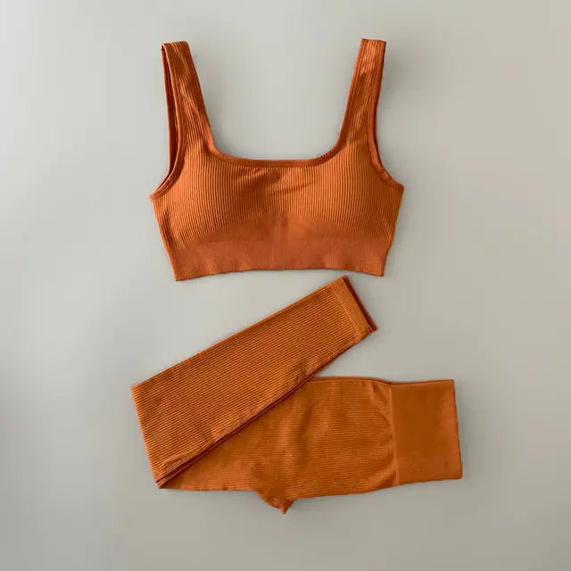 Black ribbed sports bra and matching high-waisted leggings set laid flat on a neutral gray background. The outfit is designed for activewear or athleisure, featuring a scoop neckline and wide straps on the bra.