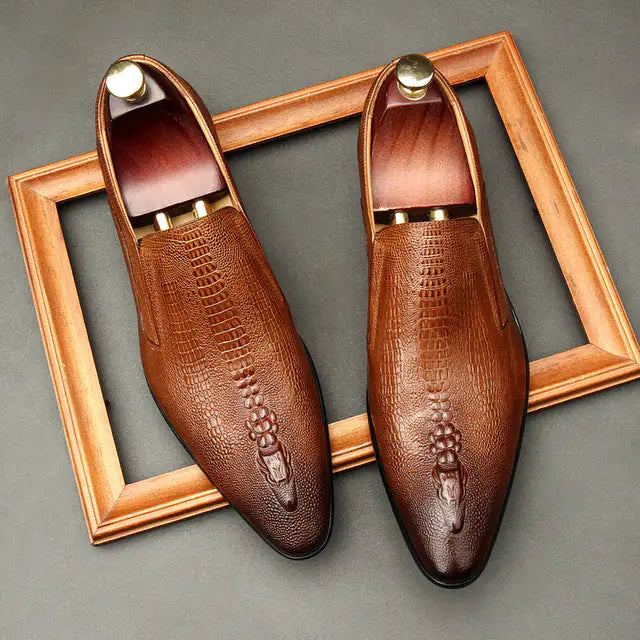 A pair of elegant burgundy leather loafers with a glossy finish. The shoes feature gold-toned metal horsebit detailing across the uppers and are displayed with wooden shoe trees inserted. They have a sleek, elongated silhouette with subtle stitching along the toe cap. The loafers are placed on a dark surface, partially framed by a wooden picture frame, emphasizing their classic and sophisticated style.