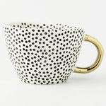 White ceramic mug covered in small black polka dots with a shiny gold handle, shown on a white background.