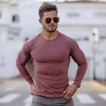 A stylish man with a fit physique is wearing a form-fitting, long-sleeved, ribbed sweater and sunglasses. He stands confidently outdoors on a street with a blurred background of white buildings. His well-groomed beard and short, styled hair complement his fashionable look, giving off a modern and sophisticated vibe