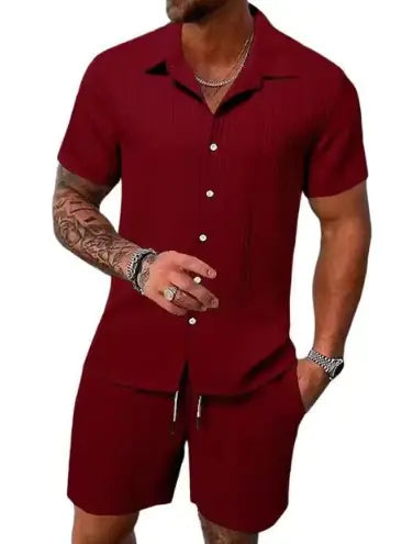 A stylish man with a muscular build is wearing a short-sleeved, button-up green shirt with matching green shorts. He is accessorized with silver rings, a wristwatch, and a layered necklace. His left arm is tattooed, and he is posing casually near a modern poolside setting with a blurred outdoor background.