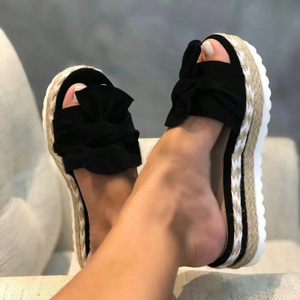 A pair of beige open-toe platform slide sandals with oversized bow details across the front. The sandals have woven espadrille-style sides with white braided trim and thick white soles, shown worn while resting on a light upholstered chair.