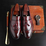 A pair of elegant burgundy leather loafers with a glossy finish. The shoes feature gold-toned metal horsebit detailing across the uppers and are displayed with wooden shoe trees inserted. They have a sleek, elongated silhouette with subtle stitching along the toe cap. The loafers are placed on a dark surface, partially framed by a wooden picture frame, emphasizing their classic and sophisticated style.