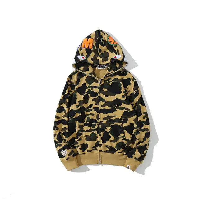 A tan and black camouflage zip-up hoodie with ribbed cuffs and hem, shown against a plain light gray background. The hood features graphic details including orange lettering and eye motifs near the top.