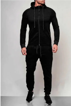 A man is wearing a stylish black tracksuit with a fitted design. The zip-up hoodie features ribbed detailing on the shoulders, black drawstrings, and zippered pockets. The matching joggers have ribbed accents on the thighs and knees, along with black drawstrings at the waist. He is also wearing black sneakers with white soles. The neutral background enhances the modern and sporty look of the outfit.