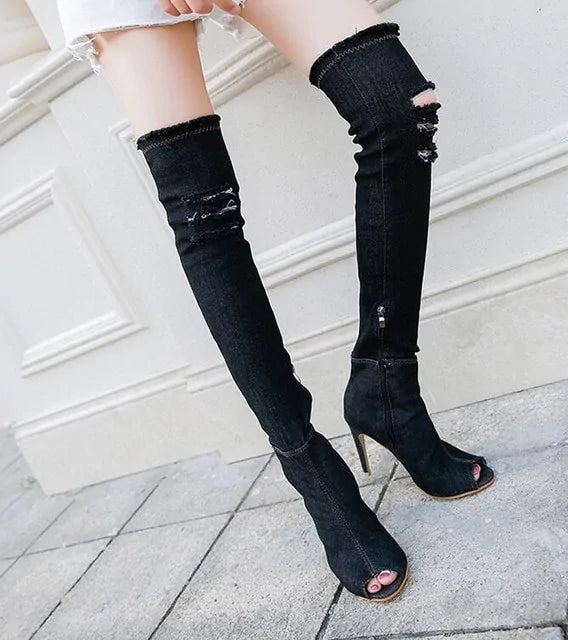A close-up view of a person wearing black, ripped, over-the-knee denim boots with peep toes and high heels, paired with frayed white shorts, standing on a stone pavement near a white wall.