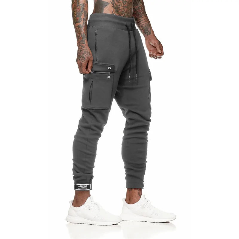 Man wearing dark gray jogger-style cargo pants with drawstring waist and multiple zip and flap pockets, paired with white sneakers, shown on a white background.