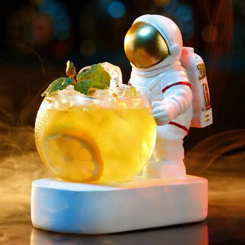 Small astronaut figurine beside a round glass of iced lemon drink with mint, sitting on a white base with warm bokeh lights in the background.