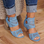 A close-up view of a person wearing stylish beige cut-out heeled sandals with ankle straps and decorative perforations, paired with cuffed blue denim jeans on a wooden floor.