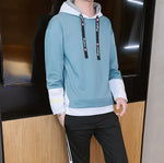 A stylish young man leans casually against a wooden wall while wearing a modern hoodie with black accents. The hoodie features black drawstrings with white text, black cuffs with embroidered details, and a red fabric tag on the left sleeve with 'LONG MA' printed on it. He is also wearing black athletic pants with white stripes on the sides. His accessories include silver rings on his fingers. The background consists of a contemporary indoor setting with a mix of wood and concrete elements.