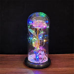 A white artificial rose inside a clear glass dome, wrapped with warm fairy lights, sitting on a round base on a wooden tabletop against a dark background.