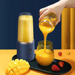 Portable blender filled with mango smoothie beside a tray as a hand pours thick yellow juice from a jar into a glass, with cut mango and strawberries on a bright yellow surface.