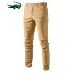 A pair of slim-fit khaki pants with a button and zipper closure, featuring a small green crocodile logo on the upper left thigh. The pants have belt loops and side pockets, with a slightly wrinkled appearance. A larger green crocodile logo is displayed in the top left corner of the image.