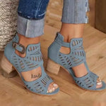 A close-up view of a person wearing stylish beige cut-out heeled sandals with ankle straps and decorative perforations, paired with cuffed blue denim jeans on a wooden floor.