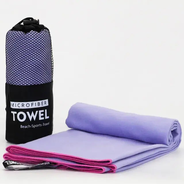 Lavender microfiber towel folded and rolled beside a black mesh carry bag labeled ‘MICROFIBER TOWEL’ with the words ‘Beach-Sports-Travel,’ shown on a white background.