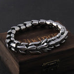 A close-up of a chunky silver-toned metal chain bracelet arranged in a circle on top of a dark wooden box, photographed against a black background.