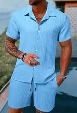 A stylish man with a muscular build is wearing a short-sleeved, button-up green shirt with matching green shorts. He is accessorized with silver rings, a wristwatch, and a layered necklace. His left arm is tattooed, and he is posing casually near a modern poolside setting with a blurred outdoor background.