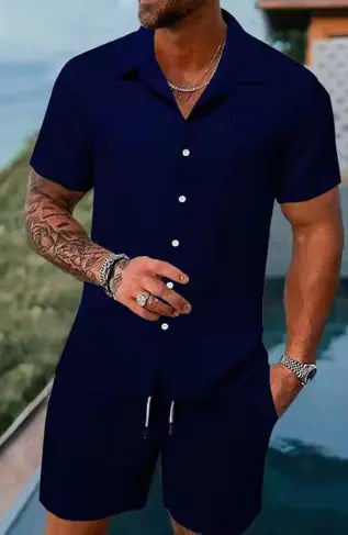 A stylish man with a muscular build is wearing a short-sleeved, button-up green shirt with matching green shorts. He is accessorized with silver rings, a wristwatch, and a layered necklace. His left arm is tattooed, and he is posing casually near a modern poolside setting with a blurred outdoor background.
