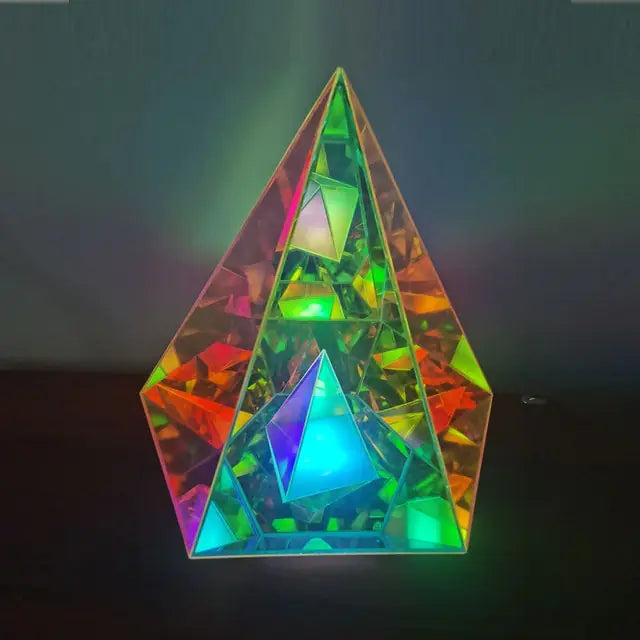 Iridescent, pyramid-shaped crystal sculpture with layered triangular facets inside, reflecting vivid green, blue, orange, and red light against a dark background.
