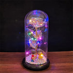 A glass dome display on a wooden surface containing a crystal-like rose wrapped with multicolored LED fairy lights, with two small figurines sitting at the base on white pebble-like beads.