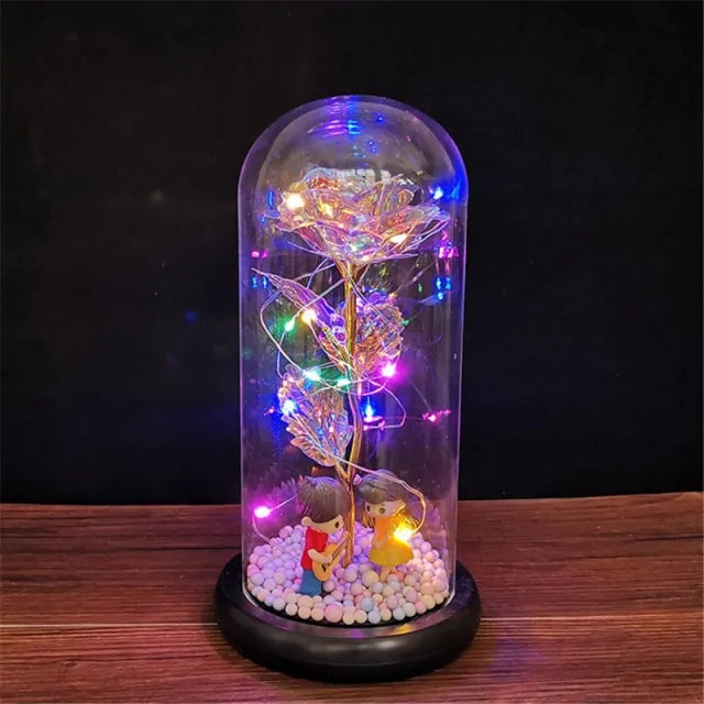 A glass dome display on a wooden surface containing a crystal-like rose wrapped with multicolored LED fairy lights, with two small figurines sitting at the base on white pebble-like beads.