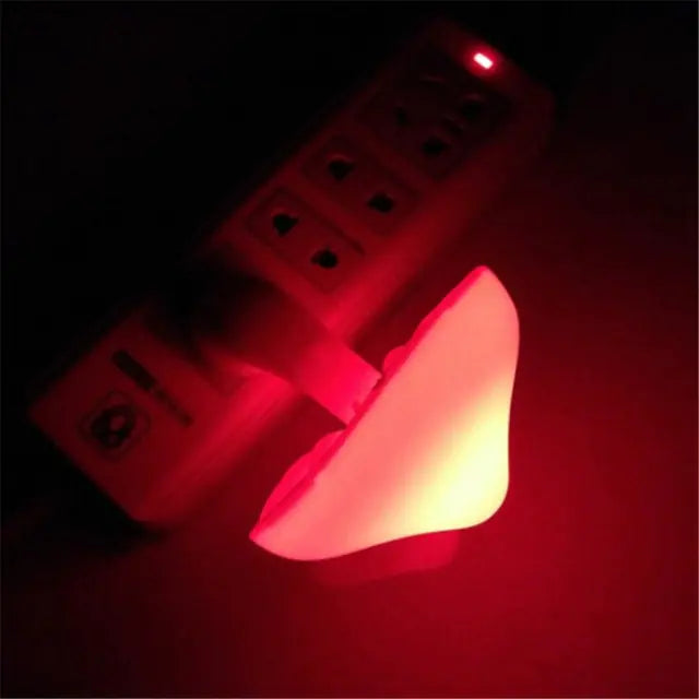 Heart-shaped red LED night light plugged into a wall socket/power strip in a dark room.