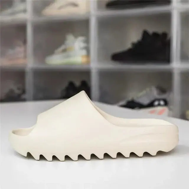 A beige Yeezy Slide with a thick, ridged sole displayed on a white surface, with a blurred background of various sneakers in transparent shoe storage boxes.