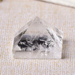 A close-up image of a clear, pyramid-shaped crystal with visible streaks or inclusions inside, resting on a textured fabric surface.
