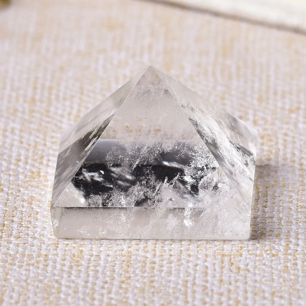 A close-up image of a clear, pyramid-shaped crystal with visible streaks or inclusions inside, resting on a textured fabric surface.