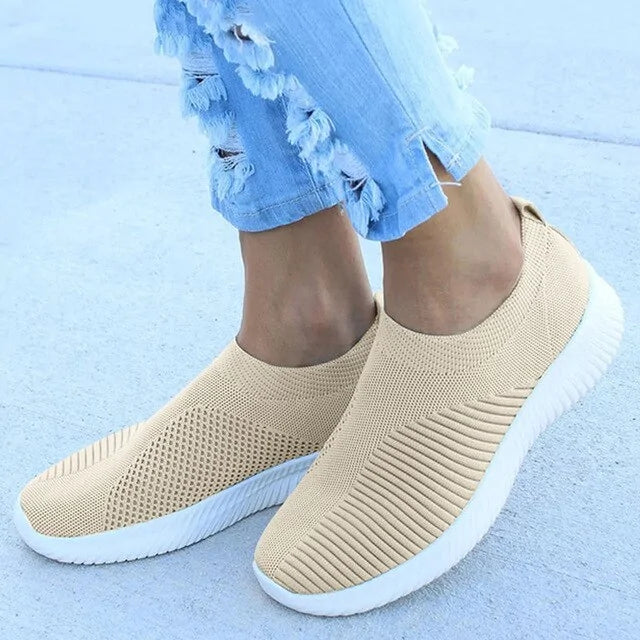 Person wearing beige slip-on knit sneakers with white ribbed soles, shown from the ankle down with light blue ripped jeans.