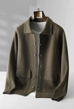 A dark olive green button-up knit cardigan with a classic collar, two front pockets with buttoned flaps, and ribbed cuffs and hem. The cardigan is layered over a white T-shirt and displayed on a wooden hanger, presenting a sophisticated and understated style.