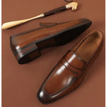 A pair of elegant brown leather loafers with a polished finish is displayed on a brown surface. The shoes feature a classic penny loafer design with a subtle decorative cutout on the strap. One shoe is positioned upright to showcase the sleek upper, while the other is laid on its side to reveal the black rubber sole. A stylish gold-handled shoehorn rests in the background
