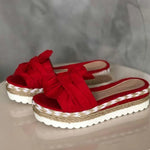 Pair of red slide sandals with oversized bow straps and braided espadrille-style platform soles with white scalloped outsoles, displayed on a tabletop.
