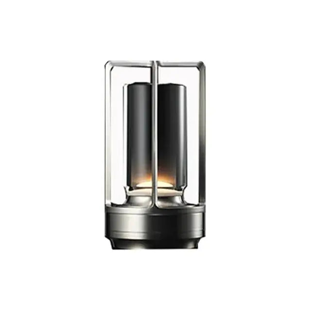 Compact modern lantern-style table lamp with a black cylindrical inner shade inside a rectangular metal frame, emitting a warm glow from the base on a white background.
