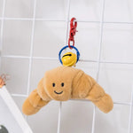 Cute plush croissant keychain with a smiley face, hanging from a colorful keyring with a yellow bell.