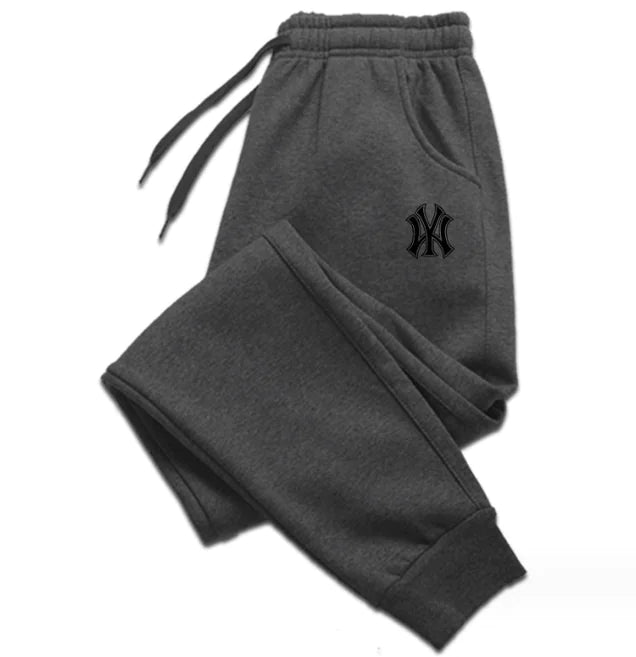 A pair of dark gray sweatpants with an elastic waistband and drawstrings, laid flat against a white background. The sweatpants feature ribbed cuffs at the ankles and side pockets. A black embroidered logo, resembling the New York Yankees emblem, is visible on the upper left thigh. The fabric appears to be soft, suggesting a comfortable and relaxed fit.