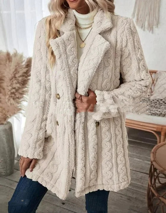 Stylish woman wearing a long red buttoned coat over a white ribbed turtleneck sweater, paired with dark blue jeans and accessorized with layered gold necklaces and a black belt. She is holding a magazine and standing in a cozy, modern room with neutral decor.
