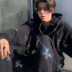 Person sitting outdoors wearing a black hoodie and matching pants covered in silver cross prints, with layered necklaces and a dark ring, lit by bright sunlight.